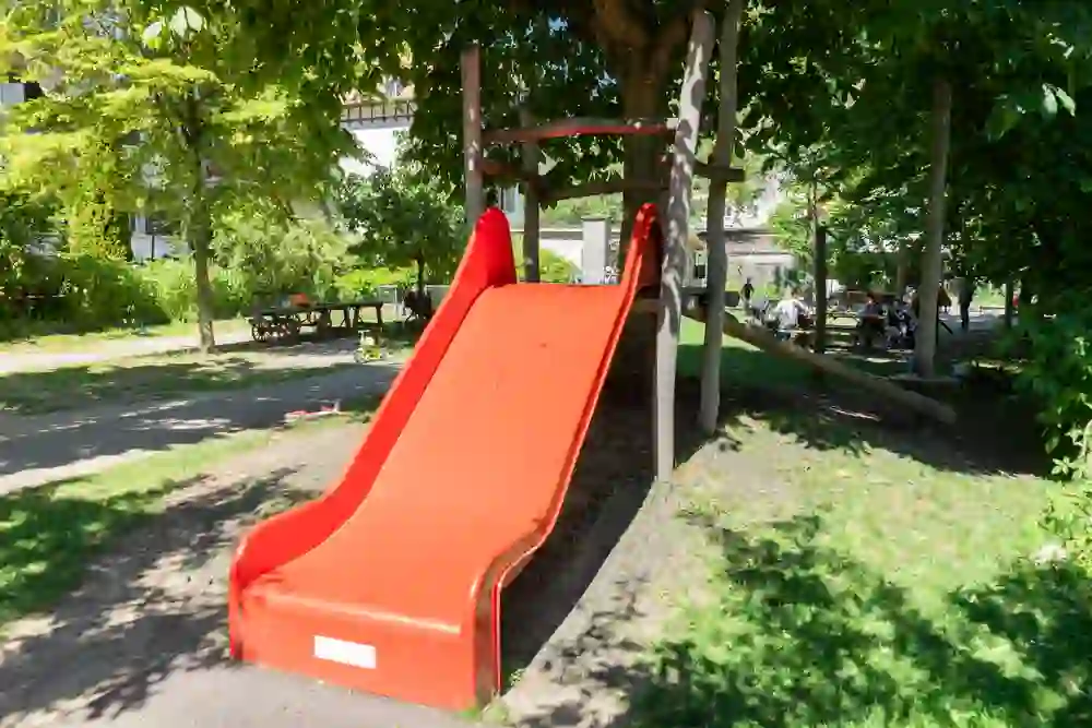Das Bild zeigt eine lebhafte, sonnige Parkszenerie mit einer leuchtend roten Rutsche, die einladend im Schatten großer Bäume steht. Im Hintergrund ist entspanntes Treiben auf Sitzbänken erkennbar, was eine friedliche und fröhliche Stimmung vermittelt.