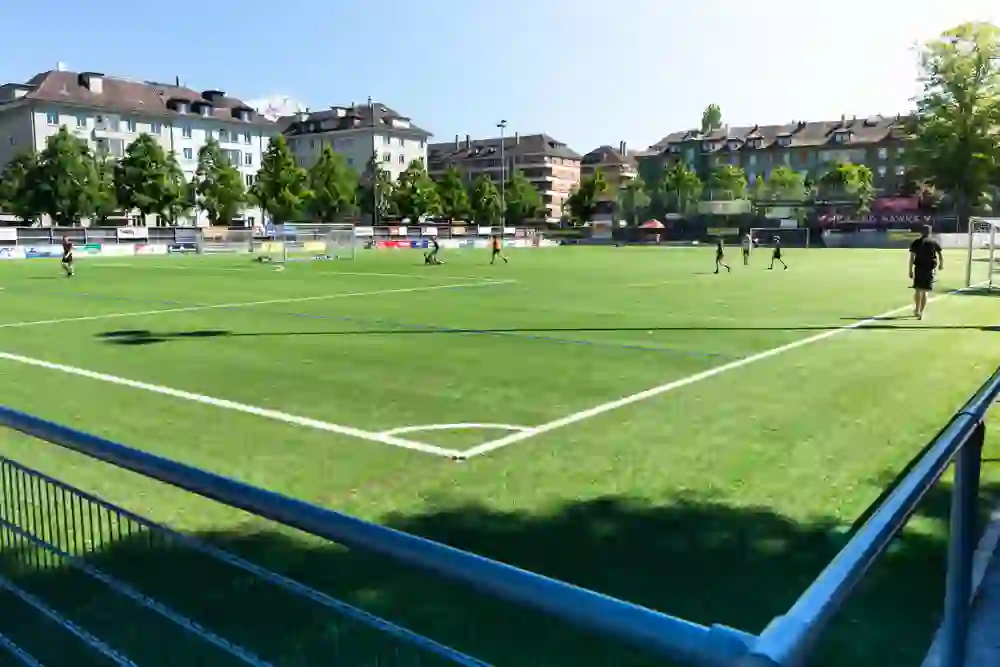 L'immagine mostra un vivace campo da calcio urbano con giocatori in azione su un'erba artificiale verde e immacolata. Circondato dall'architettura urbana, il luogo irradia energia e giovinezza, mentre il sole inonda il campo di luce intensa.