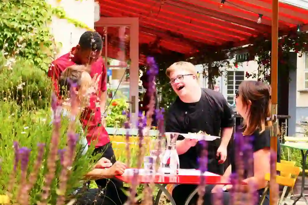 Das Bild zeigt eine lebendige Szene in einem Restaurant unter einem roten Sonnenschirm, umgeben von blühenden Pflanzen. Ein junger Mann lacht herzlich, während er sich mit Freunden unterhält, was eine fröhliche und entspannte Atmosphäre vermittelt.