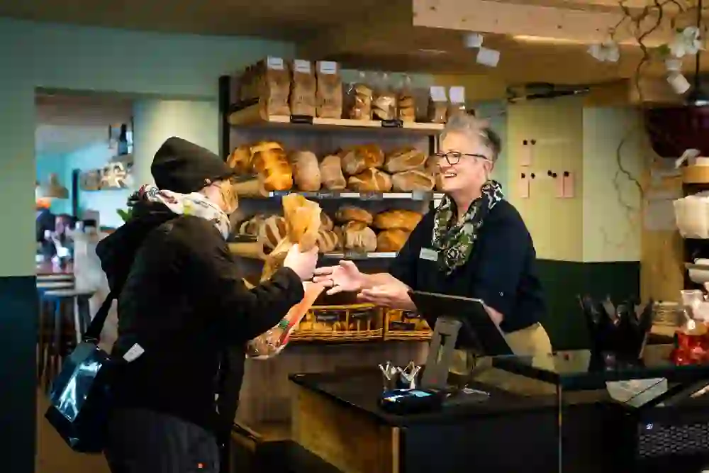 Auf dem Bild ist eine freundliche Szene in einer Bäckerei zu sehen. Es zeigt den Moment einer Kund*inneninteraktion an der Verkaufstheke, und man spürt eine herzliche, lokale Atmosphäre.