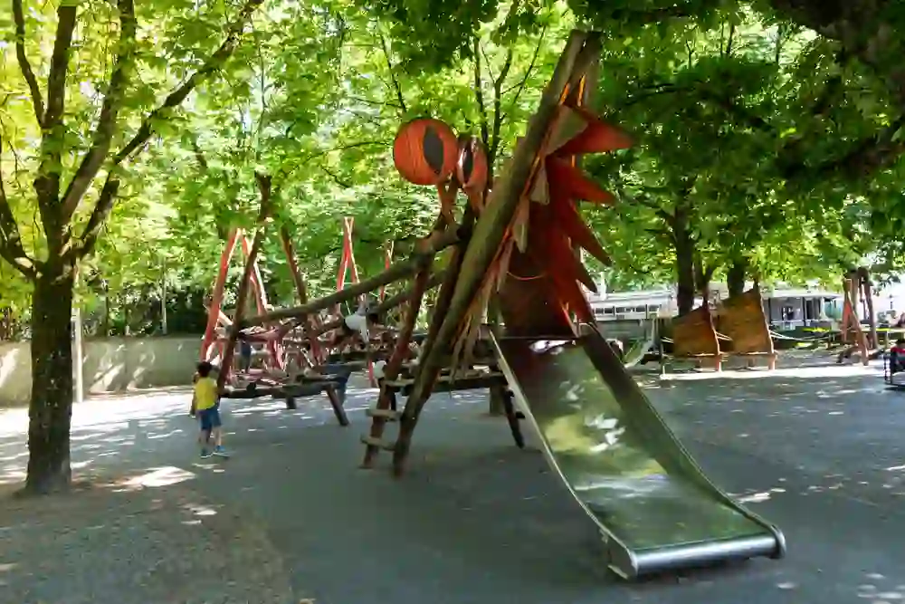 Un vivace parco giochi nel roseto di Berna, circondato da alberi rigogliosi. Il sole filtra attraverso il fogliame mentre i bambini si arrampicano e scivolano sulle attrezzature da gioco dal design unico, immersi in un mondo di divertimento e avventura.