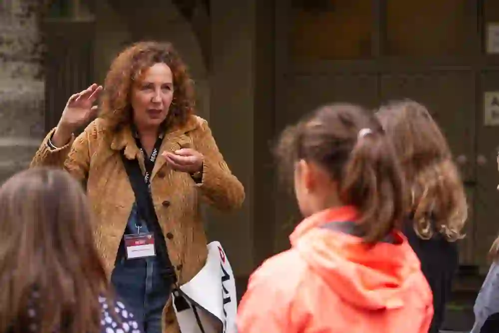 Die lebendig gestikulierende Stadtführerin erzählt eine Geschichte, die Kinder lauschen gespannt. Die Szene wirkt dynamisch und interaktiv.