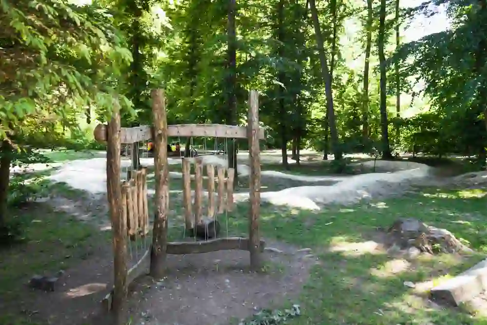 The playground in Halen near Bern invites children to play amidst lush trees and nature. A wooden sound game stands in the foreground, while children cycle on an undulating surface in the background, creating an atmosphere of discovery and joy in the green oasis.
