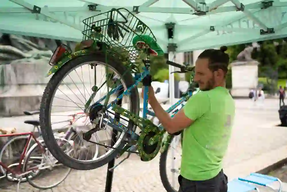 Ein Mann in einem grünen Shirt repariert sorgfältig ein bunt dekoriertes Fahrrad unter einem Zelt; sein konzentrierter Ausdruck zeigt Engagement und Fachwissen bei der Arbeit.