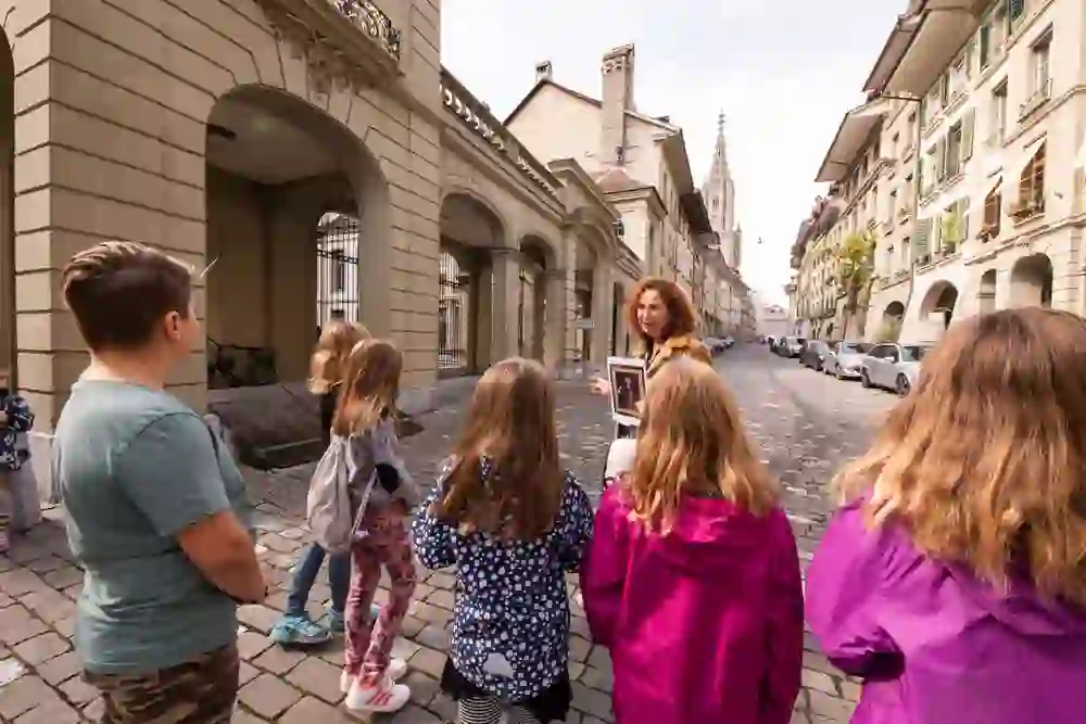 Eine Stadtführerin zeigt einer Gruppe aufmerksamer Kinder ein Bild; alle wirken interessiert und engagiert. Die Szene spielt auf einer historischen Straße mit Kopfsteinpflaster, eingefasst von traditionellen Gebäuden.