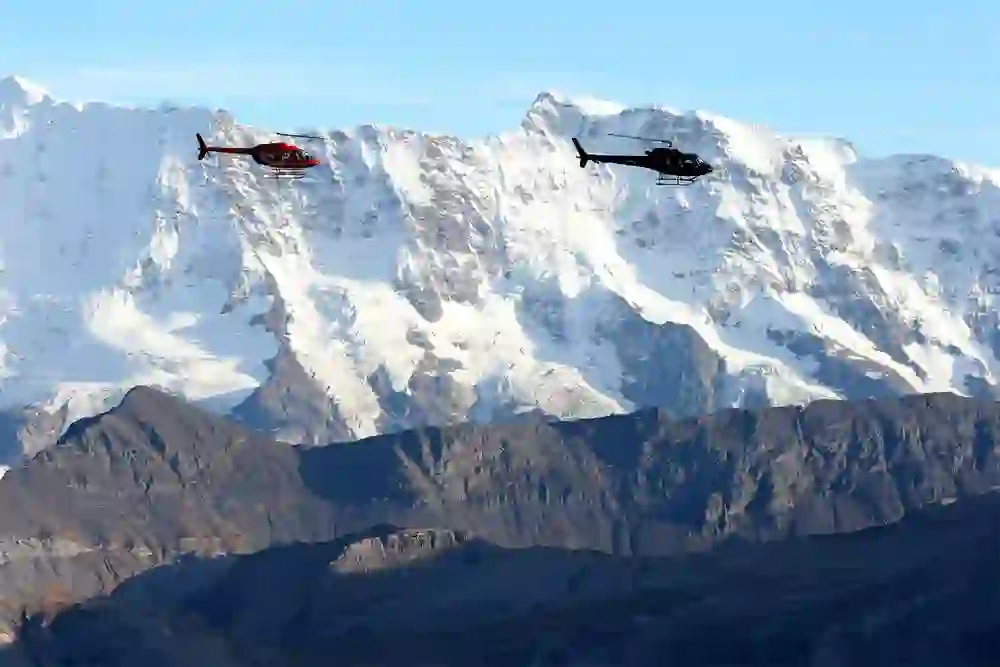 L'immagine mostra due elicotteri in volo sullo sfondo impressionante di cime alpine innevate sotto un cielo azzurro.