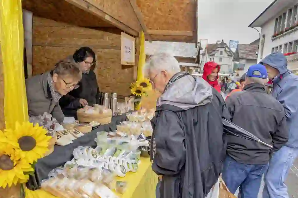 Das Bild zeigt einen belebten Käsemarkt in Huttwil, wo Menschen trotz des kühlen, grauen Wetters mit Interesse die Vielfalt an regionalen Käsespezialitäten begutachten. Die freundliche Atmosphäre und die herbstlichen Sonnenblumen verbreiten eine heimelige, gemütliche Stimmung.