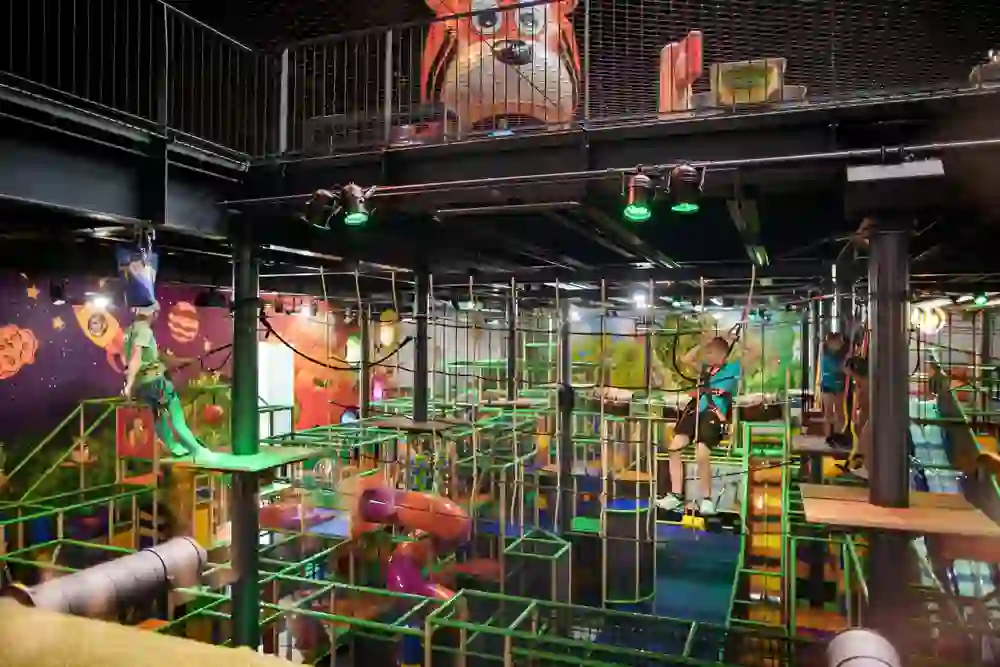 The picture shows a colourful indoor climbing park with children bravely balancing over rope bridges. The bright colours and playful designs create an adventurous atmosphere. The children radiate joy and enthusiasm as they master the obstacles.