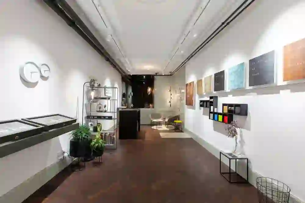 The picture shows the interior of an elegant shop, presumably a watchmaker's or jeweller's. Watches and ornate objects hang on the walls, and display cases showcase various items. The shop combines modern and classic elements, with attractive lighting that emphasises the pieces on display. The wooden floor and the calm colour scheme create a warm ambience.