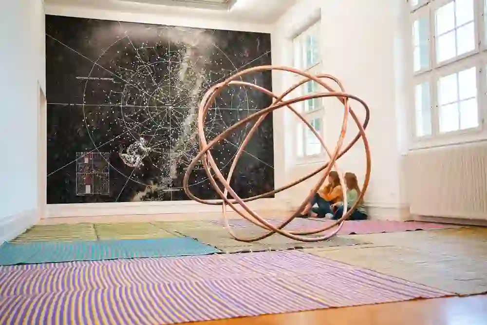 In einem hellen Raum der Kunsthalle Bern betrachten zwei Personen eine futuristische Ausstellung: eine Sternenkarte an der Wand und eine skulpturale, ringförmige Installation im Vordergrund, eingebettet in ein Meer aus bunten Teppichen.