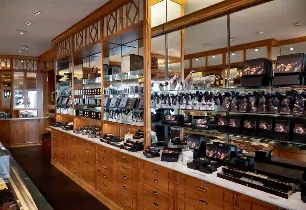 Auf dem Bild ist eine elegante Confiserie zu sehen, in der Pralinen und Schokoladen kunstvoll präsentiert werden. Die Einrichtung wirkt traditionell mit holzverkleideten Regalen und Theken sowie Glasschränken, die die edlen Süßwaren zur Schau stellen.
