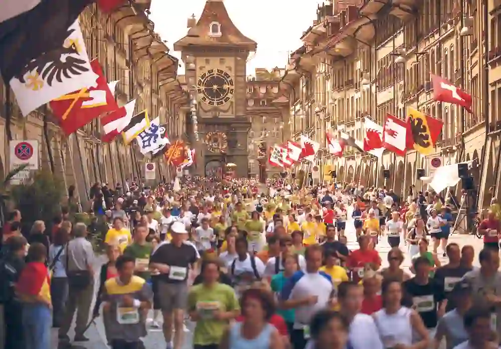 Das Foto zeigt Läuferinnen und Läufer, die am Grand Prix von Bern teilnehmen, eine Straße entlanglaufen, gesäumt von Zuschauern und Schweizer Flaggen mit der berühmten Zytglogge im Hintergrund.