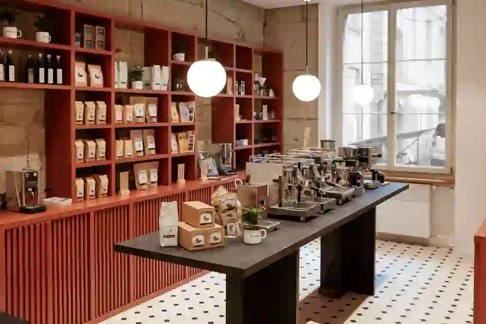 Das Foto zeigt das Innere der Gurtengasse Rösterei in Bern, ein lebhafter und stilvoller Ort. Ein rotes Regal präsentiert sorgfältig ausgewählte Kaffeesorten, während moderne Espressomaschinen auf der Theke zum Genuss einladen. Die großen Pendelleuchten verleihen dem Raum eine helle und freundliche Atmosphäre, und das geometrische Bodenmuster fügt ein dynamisches Element hinzu. Es strahlt eine moderne und einladende Wärme aus, perfekt für Kaffeeliebhaber und Freunde guten Designs.