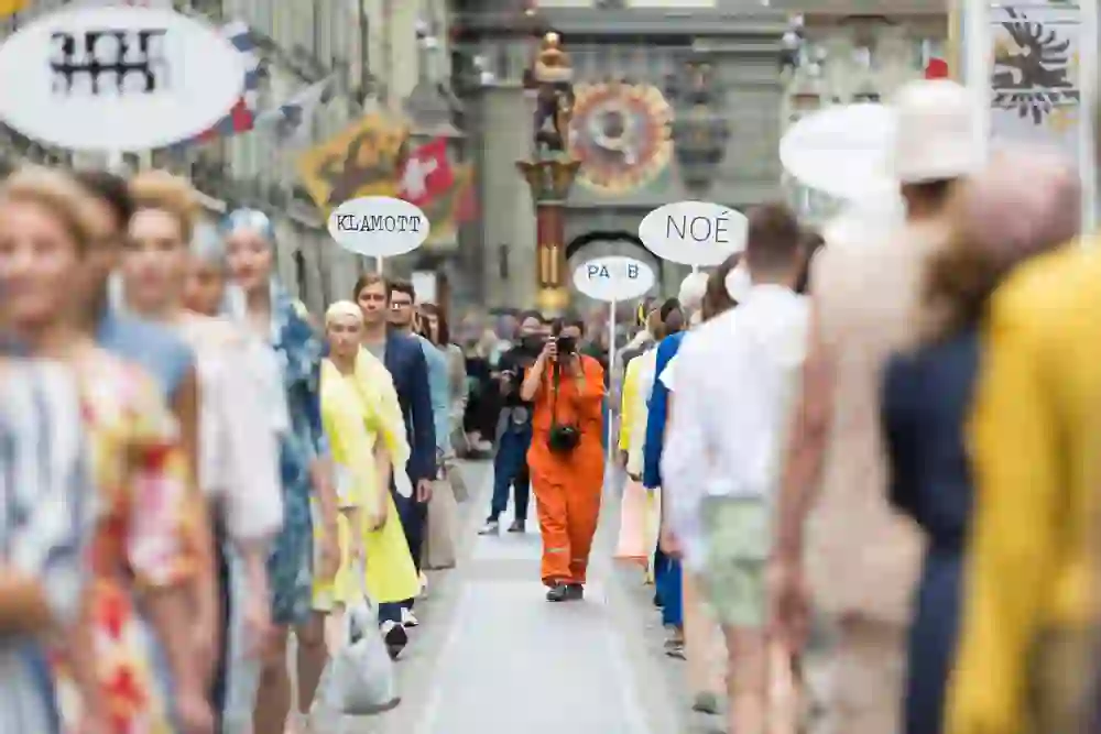 La foto mostra la vivace sfilata di moda Laufmeter per le strade di Berna. Una persona in un vistoso abito arancione, armata di macchina fotografica, si trova in primo piano, concentrata sulle modelle che passano. Le modelle indossano abiti diversi, colorati ed eleganti, che sottolineano la freschezza e la diversità della moda. Lo sfondo è pieno di spettatori e di edifici storici, che creano un ambiente affascinante e culturalmente ricco. L'atmosfera è energica e stimolante, ricca di espressione creativa e dinamismo della moda.