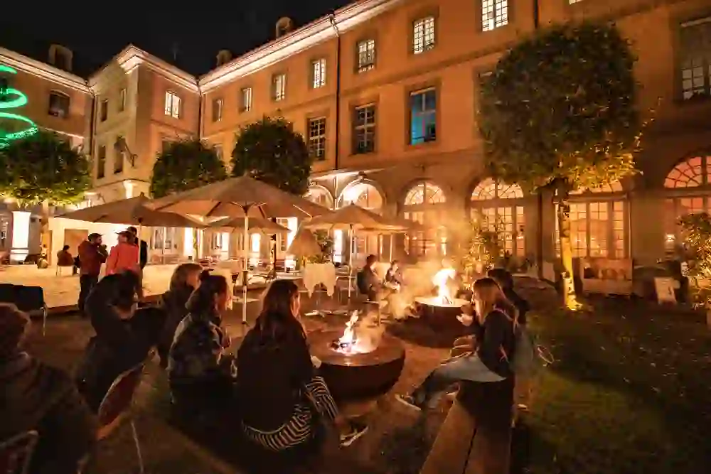 Auf dem Bild ist ein stimmungsvoller Wintermarkt im Innenhof des Berner Generationenhauses zu sehen. Menschen sitzen entspannt um Feuerstellen, genießen die Wärme und das gemütliche Ambiente unter funkelnden Lichtern in der kühlen Nachtluft.