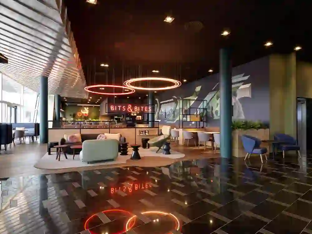 Das Foto zeigt eine moderne Hotel-Lounge des Holidays Inn Bern Westsidemit dem Namen "Bits & Bites". Auffällig sind die stilvolle Beleuchtung, farbenfrohe Möbel und geometrische Formen am Boden, die eine lebendige Atmosphäre schaffen. Es gibt Sitzbereiche mit gemütlichen Stühlen und Tischen, und die Wand ist mit einer großformatigen, abstrakten Grafik verziert. Die Beleuchtungselemente setzen warme Akzente, und die Inneneinrichtung ist ansprechend und lädt zum Verweilen ein. Die Stimmung scheint entspannt und einladend zu sein.