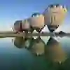 Hot air balloons with logos stand in a row on a calm lake and are reflected in the water under a clear blue sky.