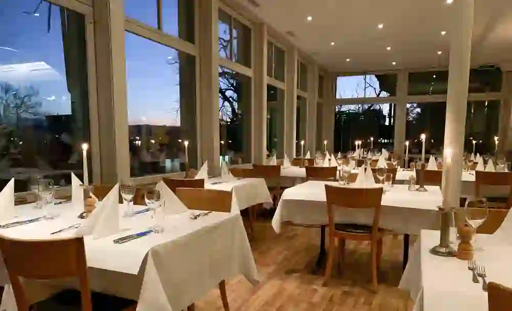 The picture shows the interior of the Veranda restaurant in the evening. Candles light up the tables and create a warm, inviting atmosphere, while the large windows offer a view of the twilight that emphasises the peaceful mood of the room.