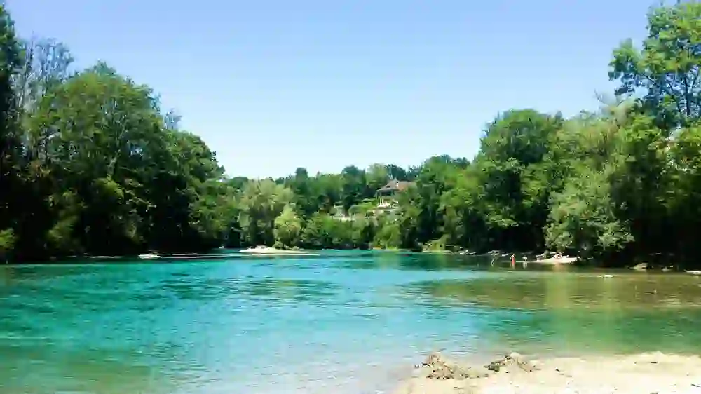 Ein ruhiger Flussabschnitt der Aare bei Bremgarten in Bern umschmeichelt eine grüne Halbinsel. Klare, blaugrüne Gewässer reflektieren das Licht, während Menschen am Ufer entspannen. Ein historisches Haus thront im Hintergrund über der friedlichen Szene, die Sommerstimmung und die Nähe zur Natur verkörpert.