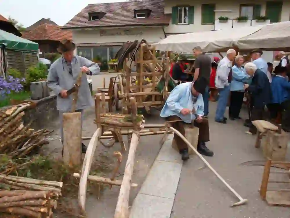L'image montre une scène d'un marché d'artisanat traditionnel. Deux hommes âgés travaillent avec des outils sur des objets en bois. L'homme à gauche écorce une branche, tandis que l'homme à droite travaille sur une pièce de bois avec un marteau. En arrière-plan, on voit d'autres personnes qui discutent ou qui regardent l'exposition. Il y a une construction en bois, peut-être un chariot, ainsi que plusieurs bancs et chaises en bois. L'événement se déroule en plein air, devant un bâtiment portant l'inscription "Blumen Rüegg".