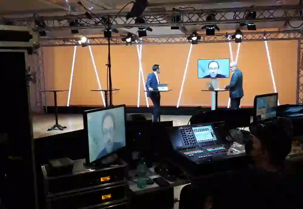 Hinter den Kulissen eines Hybrid-Events im Kongresszentrum Kreuz Bern: Techniker bedienen Ausrüstung, während Moderatoren auf Bildschirmen interaktiv präsentieren, eine moderne, dynamische Szene des digitalen Austauschs und der Vernetzung.