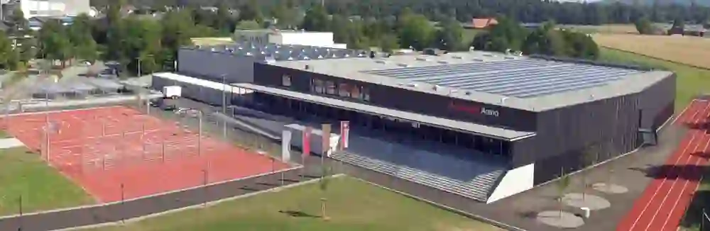 L'image montre une vue extérieure aérée de la Mobiliar Arena, un complexe sportif moderne avec de vastes terrains de sport extérieurs et une impressionnante couverture de panneaux solaires, soulignée par une atmosphère claire et énergique pour les activités et événements sportifs.