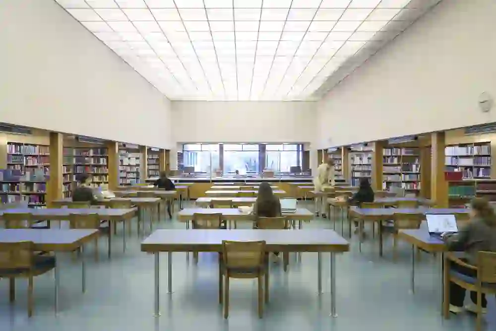 A spacious library with high ceilings, bookshelves and people studying at tables. Large windows allow daylight to flood into the room.