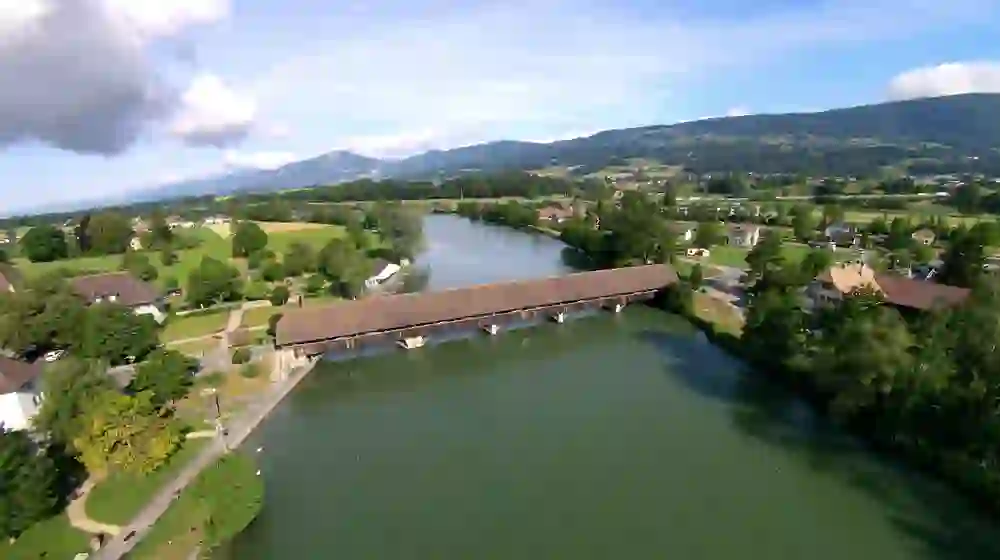 La photo montre un pittoresque pont en bois qui enjambe l'eau calme à Wangen an der Aare. Entourée de paysages verdoyants et du doux relief des collines, la photo transmet un sentiment de sérénité et d'harmonie.