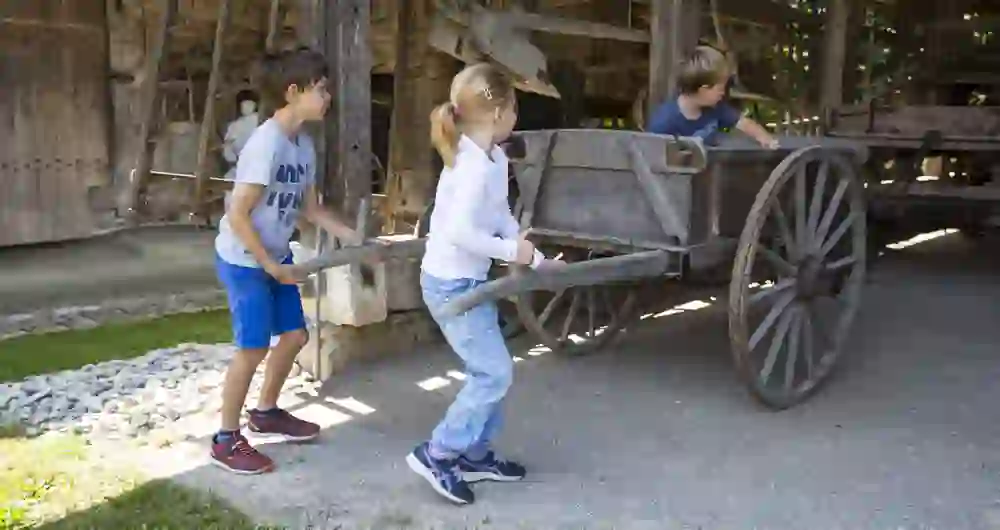 Das Foto zeigt Kinder, die im Bauernmuseum Althuus mit einem alten Holzwagen spielen. Solche interaktiven Erlebnisse können für Kinder sehr lehrreich sein, indem sie ihnen ermöglichen, Geschichte auf eine Weise zu erleben, die für sie greifbar und verständlich ist.