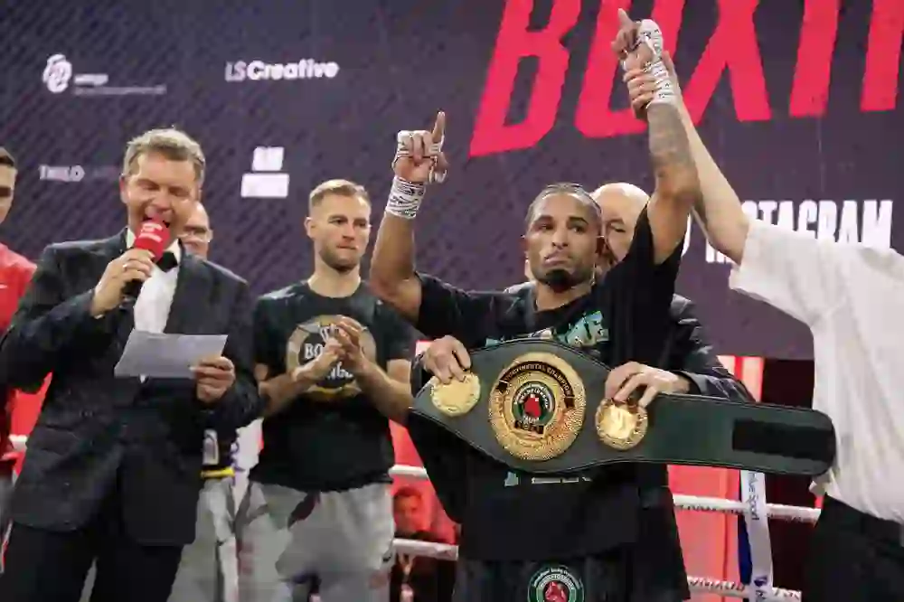 L'immagine mostra un momento di orgoglio dopo un incontro di boxe: il vincitore viene onorato con la mano alzata e la cintura di campione. Il presentatore annuncia il trionfo mentre il pubblico applaude. L'atmosfera è piena di orgoglio, sollievo ed eccitazione celebrativa.