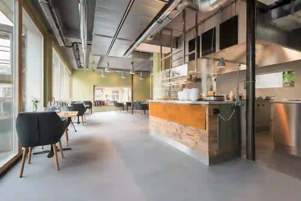 Das Bild zeigt ein modernes Restaurant mit einer offenen Küche und einem eleganten, minimalistischen Design. Die Kombination aus natürlichen Holzelementen, sanfter Beleuchtung und weitläufigen Fenstern schafft eine warme, einladende Atmosphäre, die zum Verweilen einlädt.