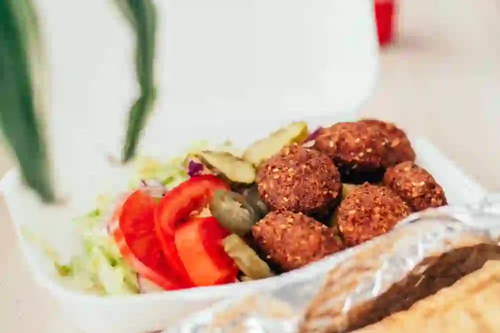 Das Bild zeigt eine appetitliche Portion Falafel mit frischem Salat, Tomaten und eingelegten Gurken, serviert in einer Takeaway-Box. Die lebendigen Farben und die frischen Zutaten vermitteln eine gesunde und genussvolle Mahlzeit.