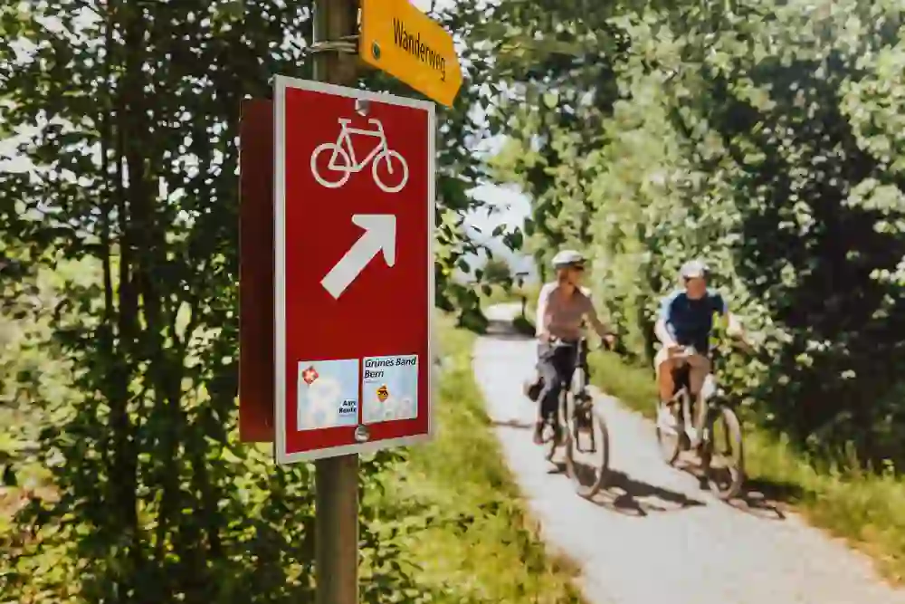 L'immagine mostra un cartello ciclistico che indica un percorso a destra. Il cartello è di colore rosso intenso, con il simbolo della bicicletta e la freccia bianchi. Sotto di esso sono presenti altri cartelli con le scritte "Grünes Band Bern" e "Aare Route". Sullo sfondo, due persone stanno percorrendo in bicicletta un sentiero ombreggiato e circondato da alberi e vegetazione. Sembra una giornata soleggiata e piacevole per una passeggiata in bicicletta.