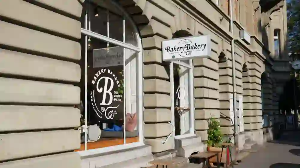 The picture shows the exterior of a charming vegan bakery on a sunny day. The façade shines with clear, inviting windows and cosy seating in front of the shop, creating a friendly and relaxed atmosphere.