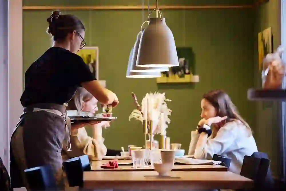 In einem gemütlichen Café mit warmem Licht serviert eine Kellnerin Speisen an einen Tisch. Zwei Gäste unterhalten sich entspannt. Die Atmosphäre wirkt freundlich und einladend, perfekt für entspannte Gespräche.