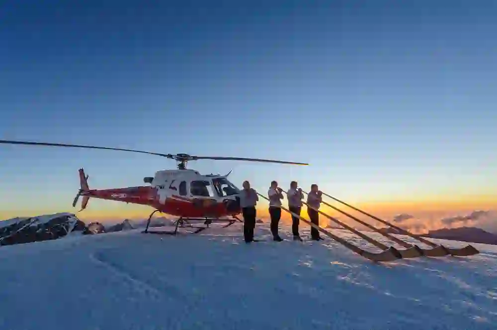 Das Bild zeigt vier Personen, die Alphörner blasen, neben einem roten Helikopter auf einem verschneiten Berggipfel bei Sonnenuntergang. Die letzten Sonnenstrahlen tauchen den Himmel in ein warmes Farbenspiel aus Orange und Blau.