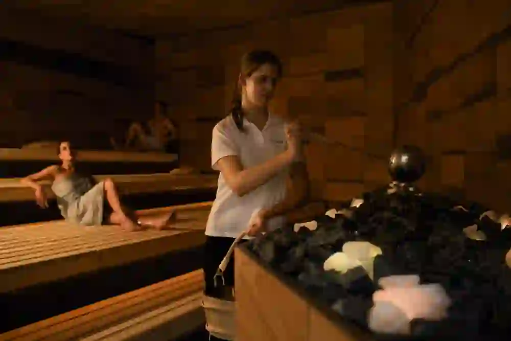 Eine Saunameisterin im Bernaaqua giesst konzentriert Wasser auf heiße Steine, während die Gäste in der wohligen Wärme der Sauna entspannen und in ruhiger Atmosphäre auf den Bänken liegen.