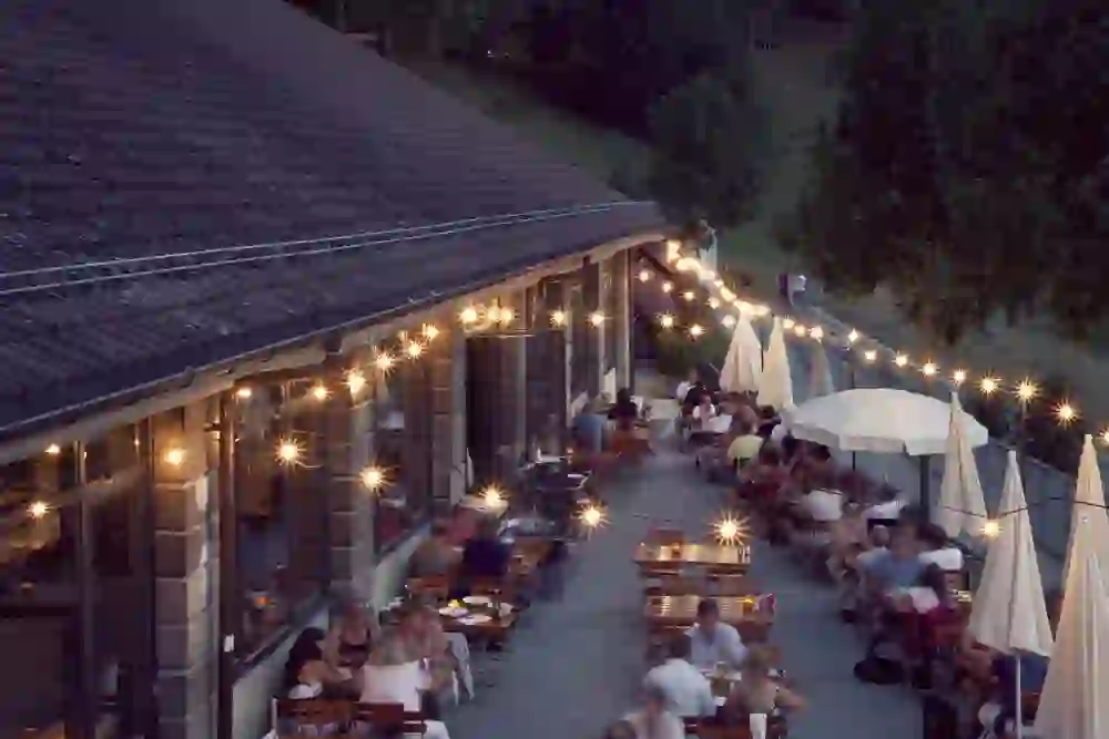The picture shows the outdoor ambience of the Altes Tramdepot restaurant at dusk. Warm lights shine over the terrace, where people sit and relax while chatting and eating. The atmosphere is cosy and inviting.
