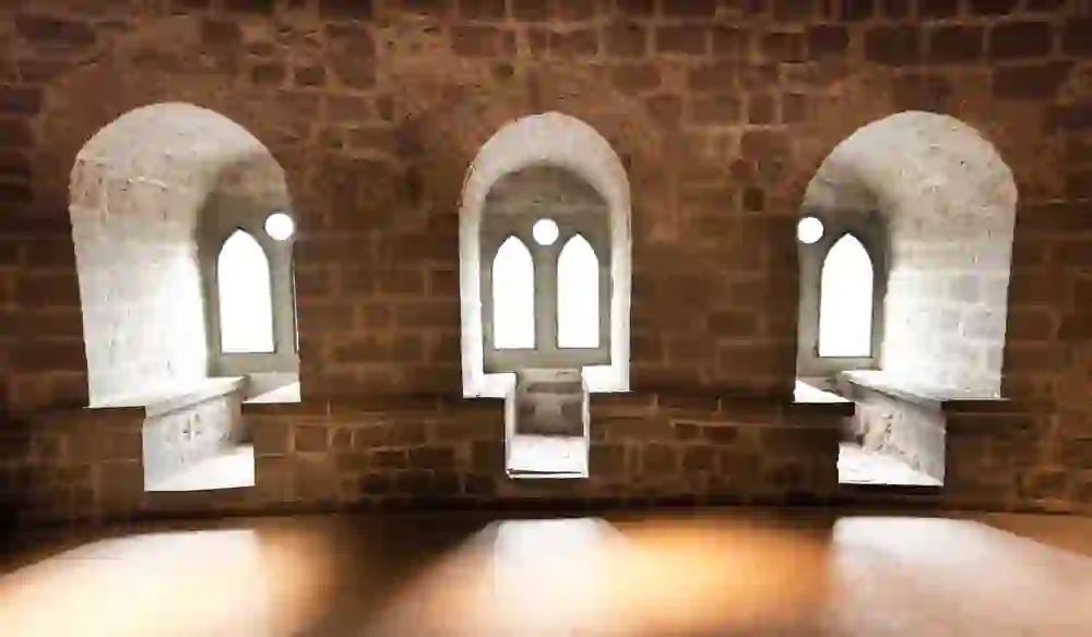 Das Bild zeigt drei schmale, hohe Fenster mit spitzbogigem Abschluss, eingebettet in das dicke Mauerwerk eines Schlosses. Das warme, diffuse Licht, das durch die Fenster hereinfällt, erzeugt eine ruhige und besinnliche Atmosphäre, die die historische Bedeutung und das Alter des Gebäudes – Schloss Laupen – unterstreicht.