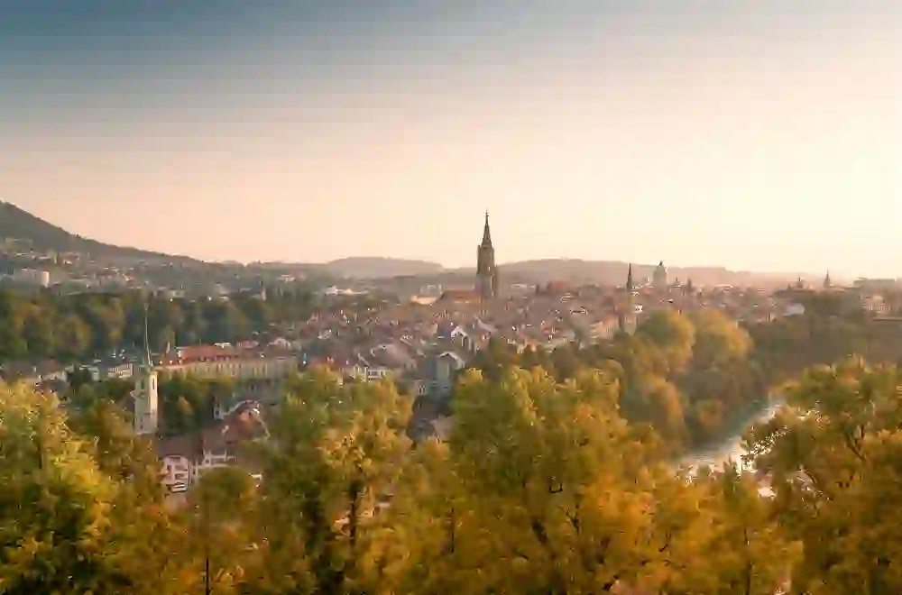 Ein sanftes Herbstlicht umarmt die Stadt Bern, wobei die charakteristische Silhouette des Münsters sich elegant über die bunten Bäume und die ruhige Aare erhebt, ein Gefühl von Frieden und zeitloser Schönheit vermittelnd.