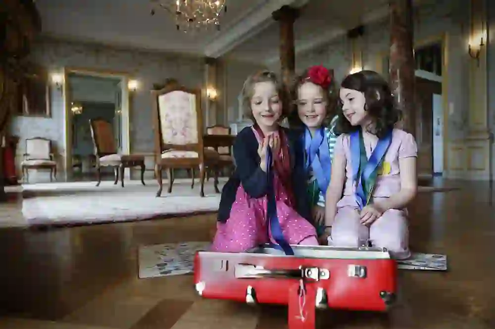 Three little girls, adorned with sashes and medals, laugh and play together on the glittering parquet floor of an opulent palace hall. The joy and amazement on their faces are in harmony with the magic of their magnificent surroundings.