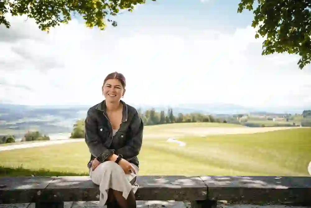 Tatjana Schär, eine junge Frau mit einem freudigen Lächeln, sitzt entspannt auf einer Bank, eingebettet in die friedliche Weite einer grünen Landschaft.
