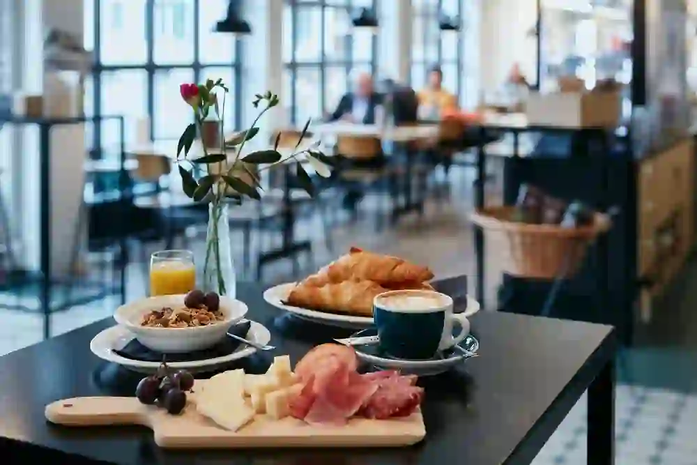 Auf dem Bild ist ein Frühstücksarrangement der Röstereie Kaffee und Bar zu sehen. Im Vordergrund ein Tisch mit einer Auswahl an Käse und Wurst auf einem Holzbrett, einer Schale Müsli, einem Croissant und einem Cappuccino. Im Hintergrund sind Gäste und das Caféinterieur unscharf zu erkennen. Ein Glas Orangensaft und eine Vase mit Blumen vervollständigen die Szene.