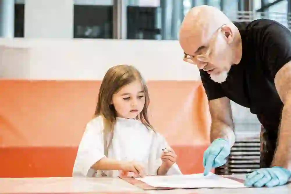A girl and an adult in a black shirt, who is supporting her in a creative activity, immerse themselves in a work of art, their shared focus radiating intimacy and pedagogical care.