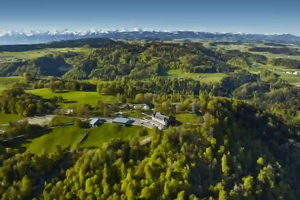 Eine Drohnenaufnahme offenbart den malerischen Gurtenpark, umrahmt von saftig grünen Wäldern und leuchtenden Feldern. Im Hintergrund erhebt sich das majestätische Alpenpanorama, das die Szenerie mit einer atemberaubenden Kulisse krönt.
