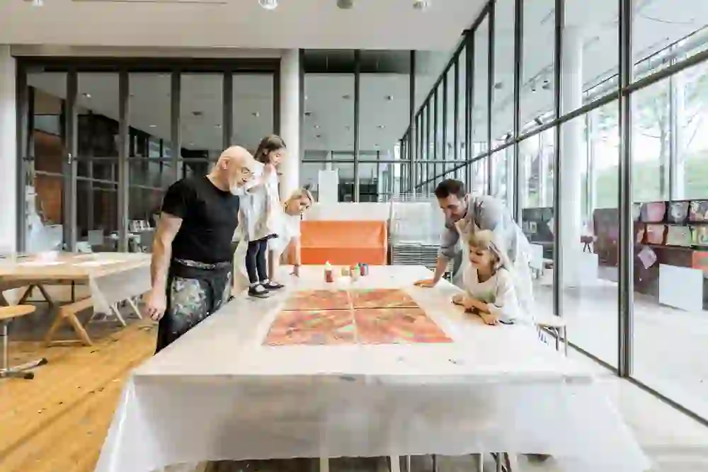 Erwachsene und Kinder betrachten gemeinsam Kunstwerke auf einem Tisch in einem hellen Raum. Die konzentrierten Gesichter und entspannte Atmosphäre vermitteln einen Eindruck von Kreativität und Lernfreude.