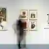 A blurred person walks past a gallery wall on which various colourful collages and works of art are displayed in frames.
