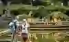 The picture shows a public performance by dancers positioned in synchronised poses on yellow platforms in the Rose Garden in Bern. The dancers wear uniform, earth-coloured costumes and are obviously performing a well-choreographed sequence. In the foreground, two spectators - an elderly couple with rucksacks - watch the performance. The tranquil pond in front of them reflects the scene, emphasised by the green of the surrounding trees and bushes. This idyllic, almost surreal setting creates an enchanted atmosphere that captures both the tranquillity of the location and the dynamism of the performance.