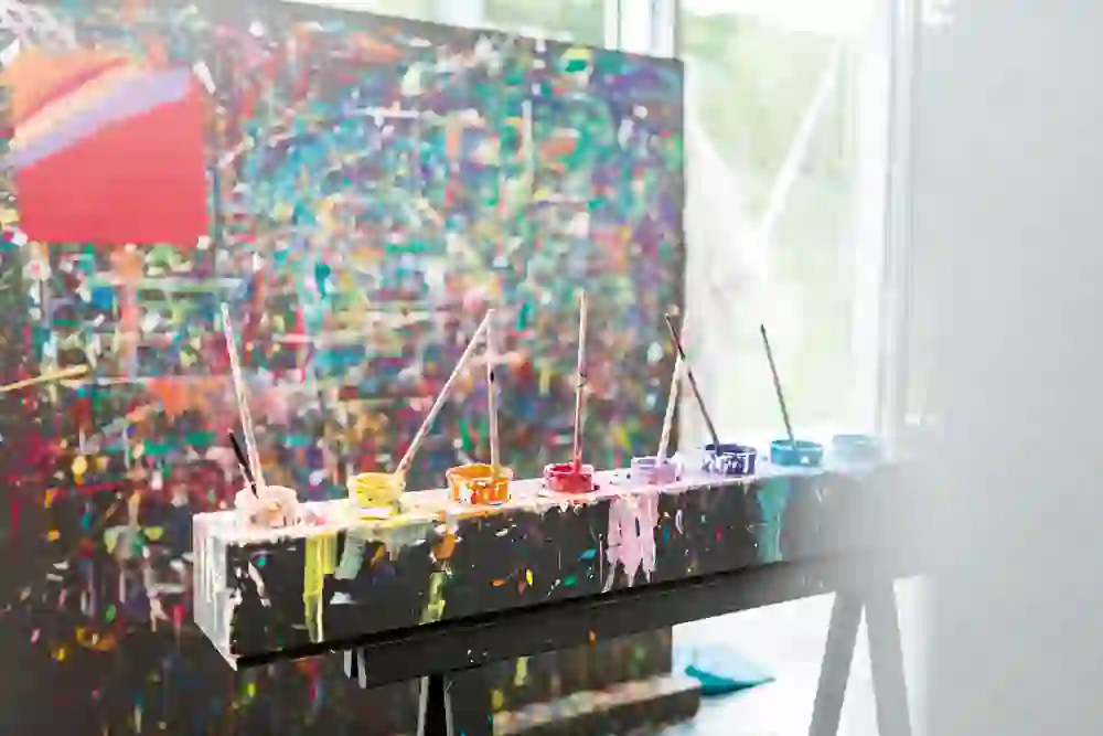 Ein explosiver Ausdruck von Kreativität: Farbgefüllte Pinsel ruhen auf einer Staffelei vor einem lebendigen, farbenfrohen Gemälde, das Licht und Inspiration ausstrahlt.