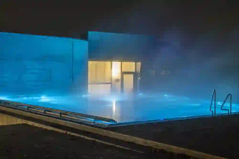 Ein erleuchteter Aussenpool strahlt in mystischem Blau in der Nacht, umgeben von moderner Architektur und Nebelschwaden, ein Bild der Stille und Entspannung im Wellnessbereich AquArenA.